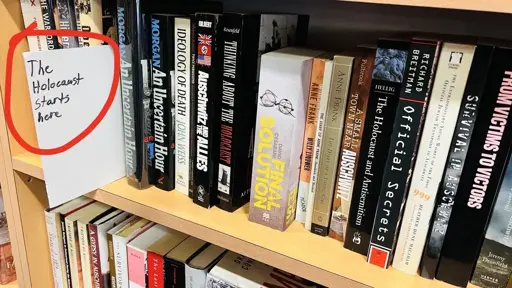 Books on a shelf with a hand written sign, presumably by book store staff, that says “The Holocaust starts here”.
